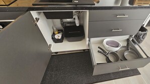 Una cocina con encimera negra, pileta blanca y un cajón con utensilios de cocina.