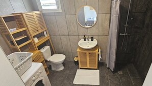 Un baño pequeño con inodoro, lavabo y ducha. Hay estantes de madera y una lavadora.