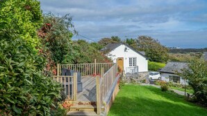 A white cottage with a wooden deck and a small garden, surrounded by lush greenery.