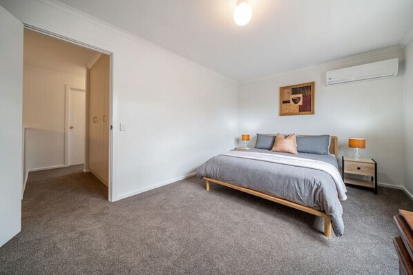 Un dormitorio con cama, mesitas de noche, aire acondicionado instalado en la pared y una puerta que da a otra habitación.