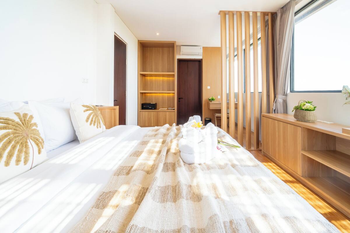 Une chambre d’hôtel moderne avec un grand lit, des étagères en bois, un petit bureau et une fenêtre donnant sur un beau paysage.