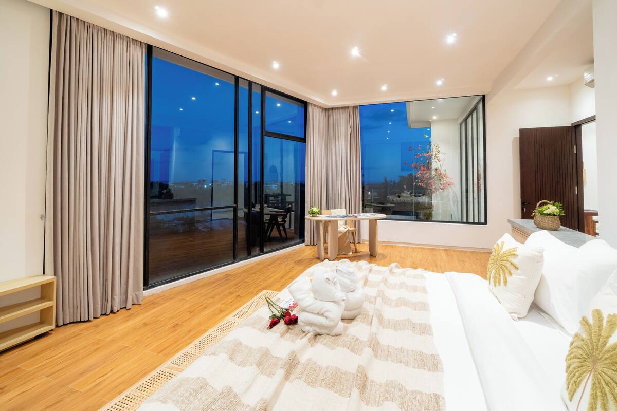 Une chambre moderne avec une grande fenêtre, un canapé, une table de chevet et une vue sur le paysage urbain au crépuscule.