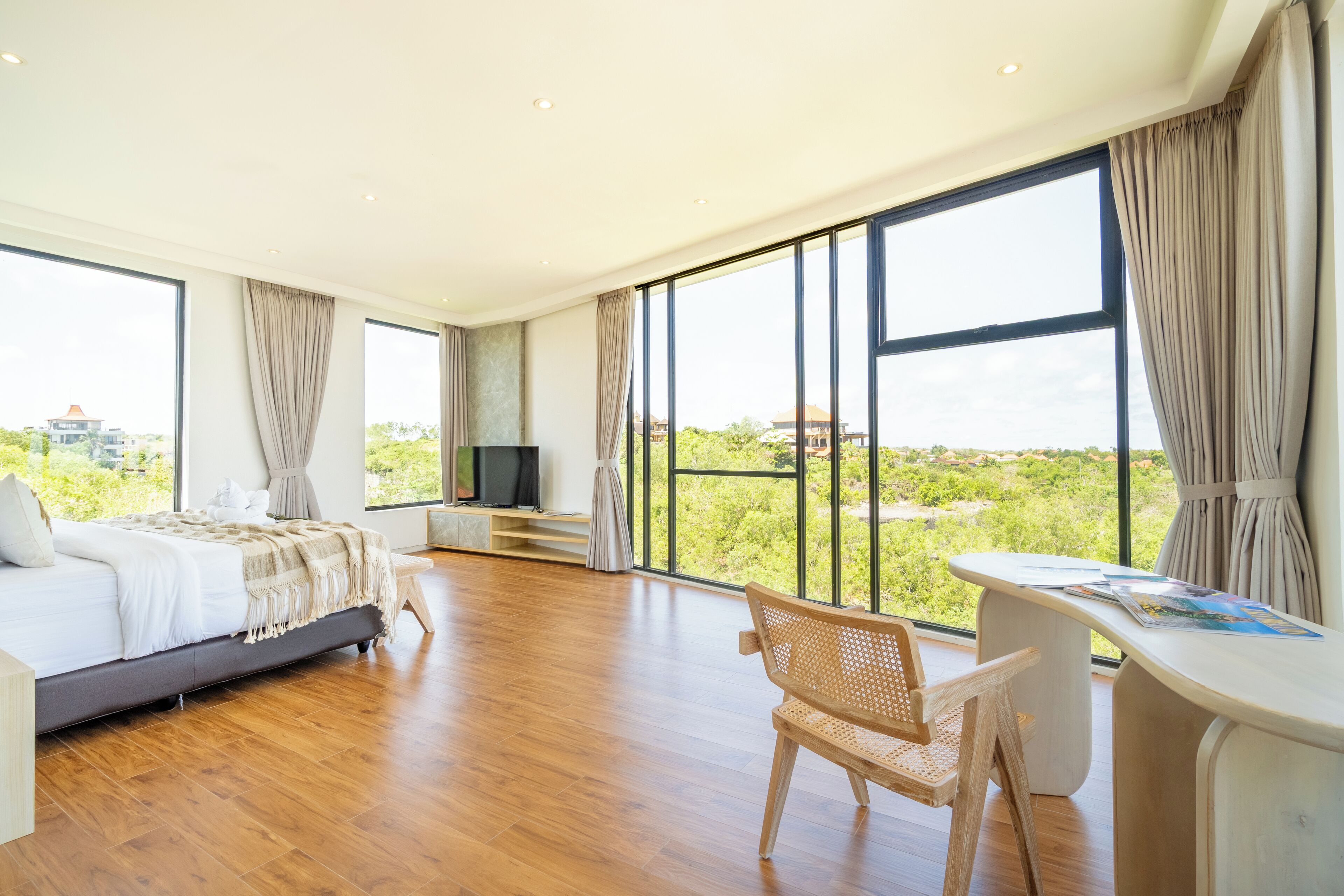 A spacious bedroom with a large bed, a desk, and a view of greenery through large windows.