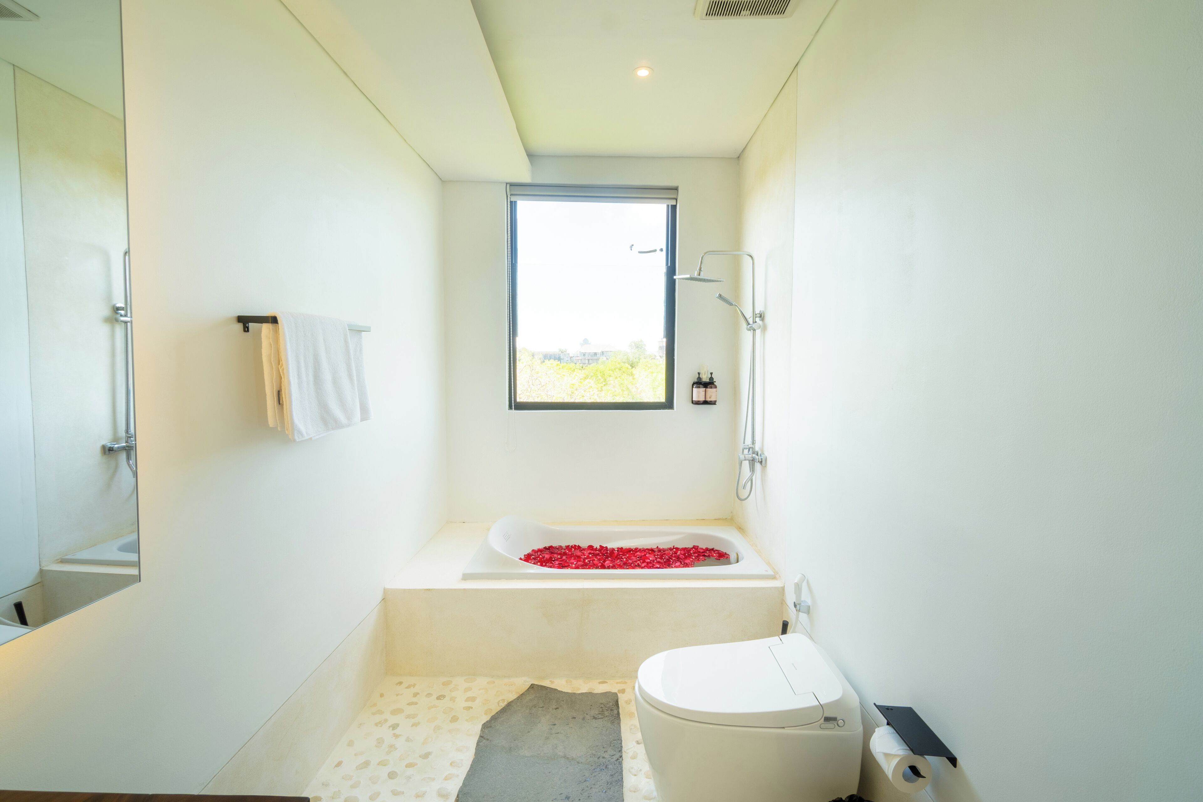 Un baño moderno con una bañera llena de pétalos de rosa roja, un inodoro y una ventana que deja entrar luz natural.