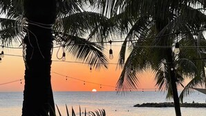 A beach scene with palm trees, string lights, and sun setting over the ocean. - Upscale Blue Bay Golf & Beach Suite – Gated Resort – 5 Min Walk to Beach (Sint Michiel)