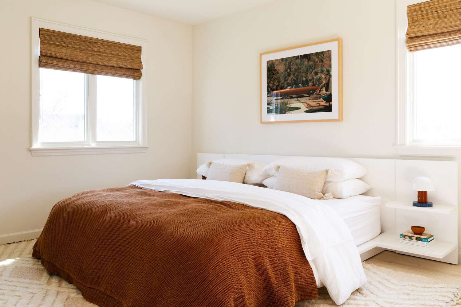A bedroom with a large bed, white bedding, a brown throw blanket, a nightstand with a lamp, and a framed picture on the wall.