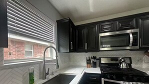 Une cuisine moderne avec des meubles noirs, des appareils en acier inoxydable, une fenêtre avec des stores et une vue sur un bâtiment en briques.