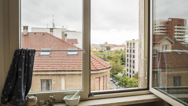 Ein Zimmer mit Fenster und Blick auf eine Stadtlandschaft, mit einem Ziegeldach, einem Gebäude mit vielen Fenstern sowie einer Ansicht auf weitere Gebäude und Grünflächen.