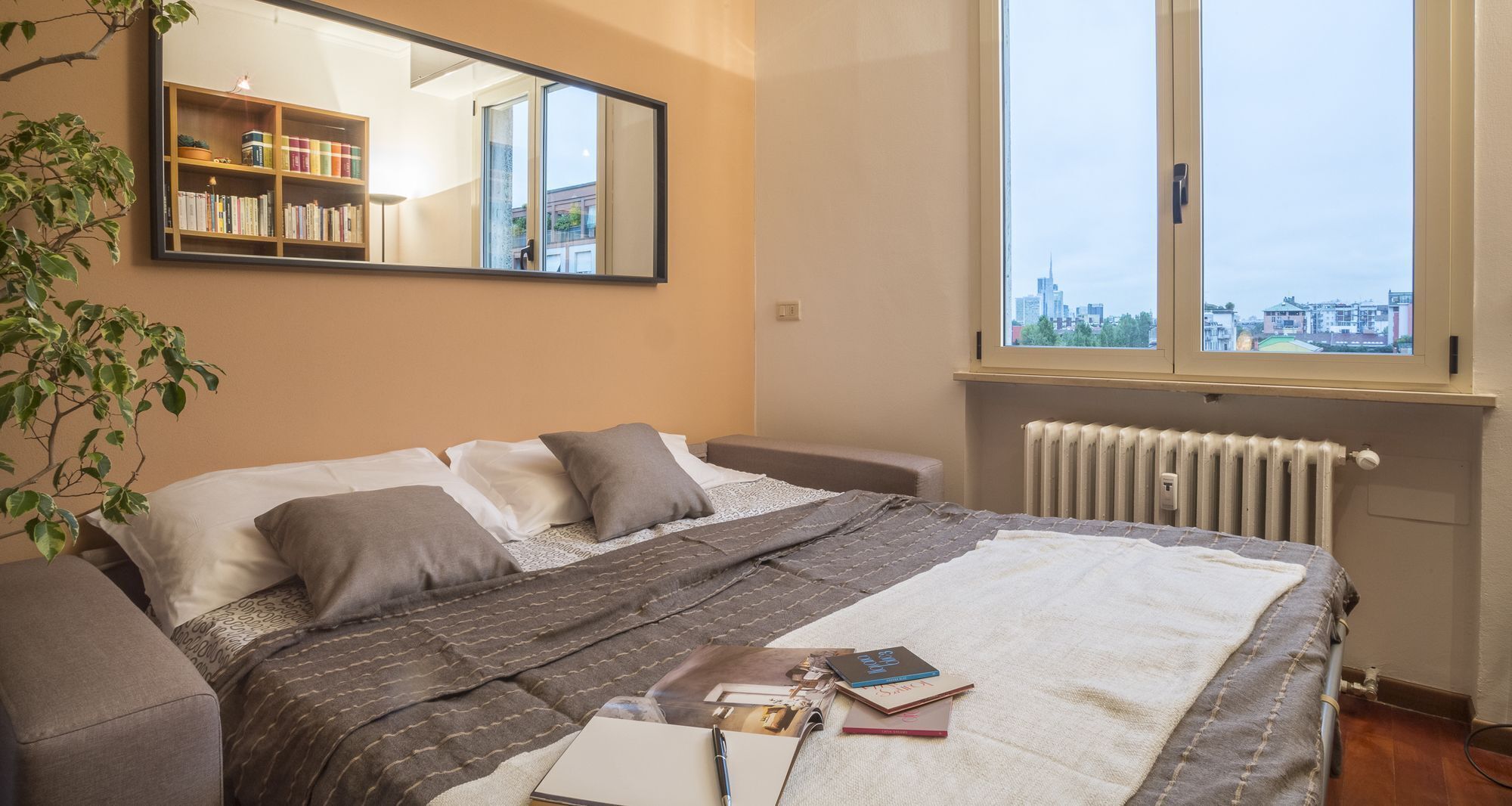 Une chambre à coucher comprenant un lit, une table de chevet, un radiateur, une fenêtre donnant sur la ville et une bibliothèque.