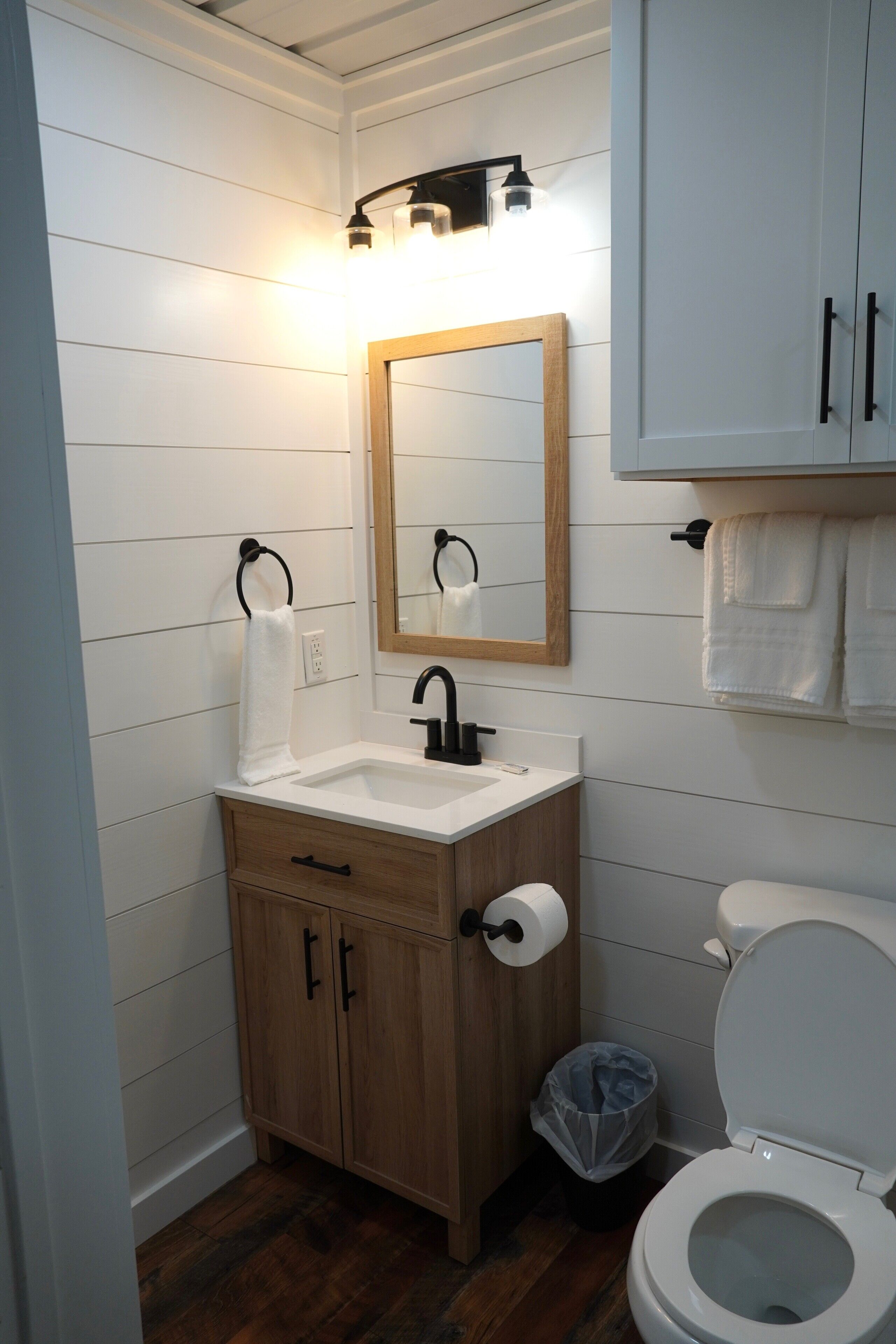 Et badeværelse med et træbord, en hvid vask, en sort vandhane og et spejl over vasken. Der er et hvidt toilet ved siden af bordet og en skraldespand foran toilettet.