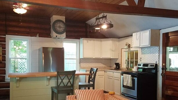 Interior acogedor de una cabaña con techo de madera, un sofá a rayas, una mesa de comedor con sillas, una zona de cocina con estufa y refrigerador, y un ventanal con vista a la vegetación.