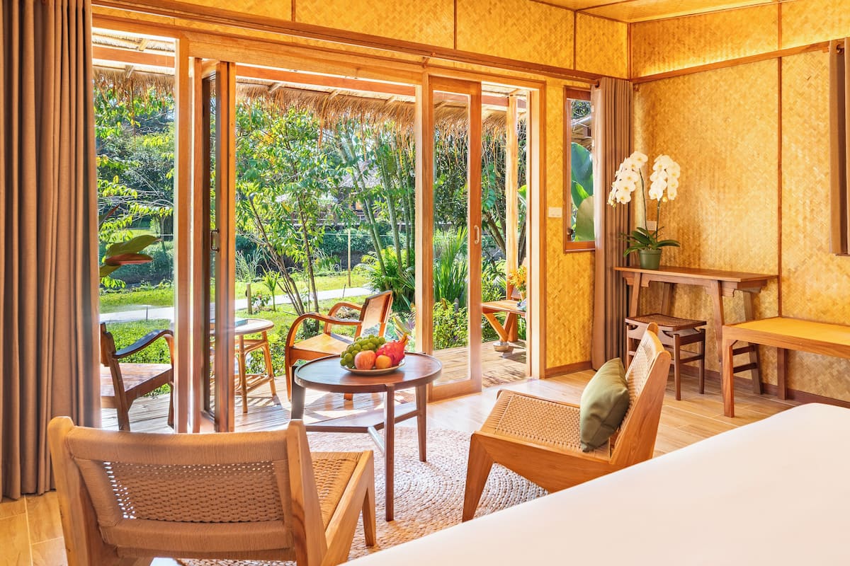 A room with a bed, a dining table with chairs, a small table with fruit, and a view of the garden through large glass doors.