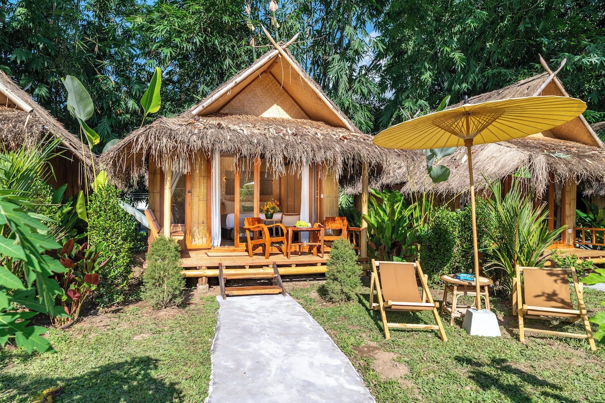 A bungalow-style accommodation with thatched roofs, wooden furniture, and a concrete pathway leading to the entrance.