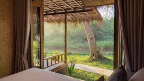 Une chambre avec un lit, des rideaux et une vue sur un paysage naturel comprenant un arbre et de l’eau.