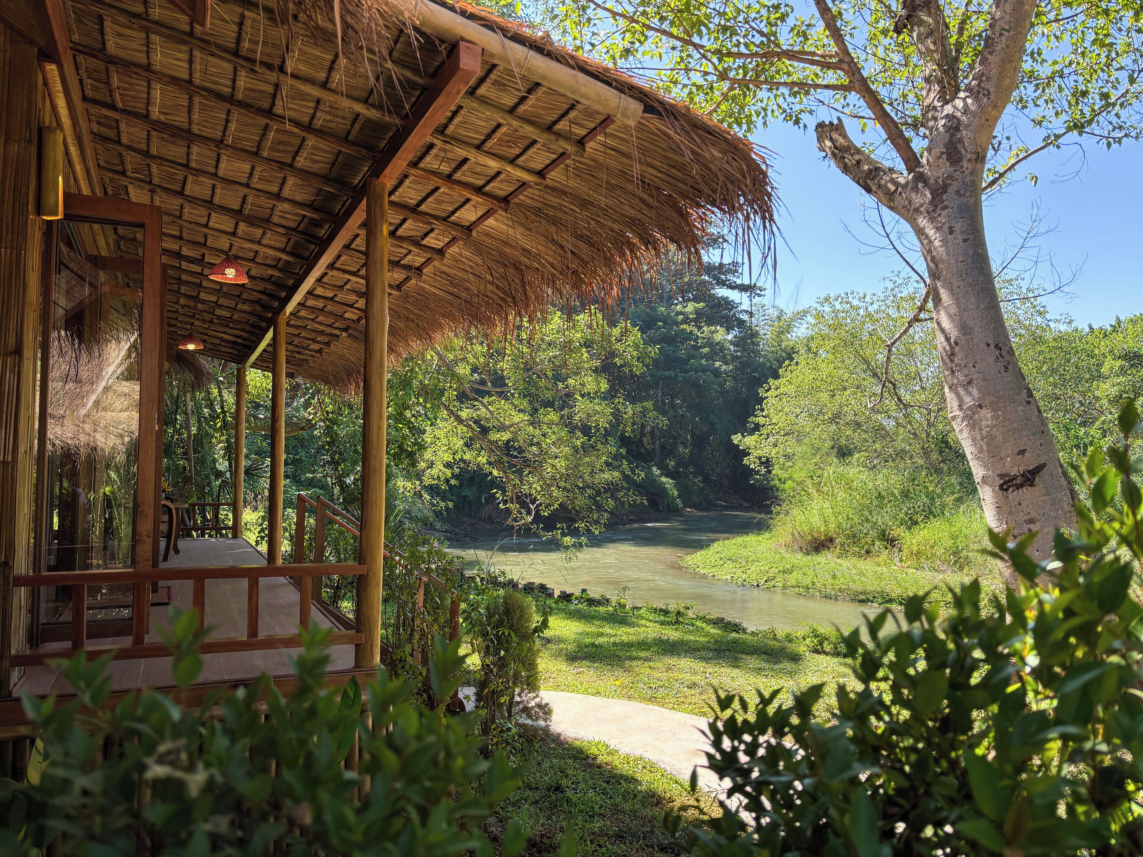A rustic wooden structure with a thatched roof, surrounded by lush greenery and a river.