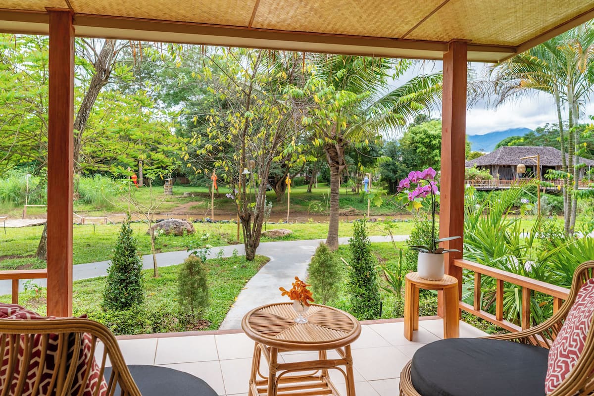 A covered outdoor area with wicker furniture, a small table with a vase, and a view of a lush garden and distant buildings.