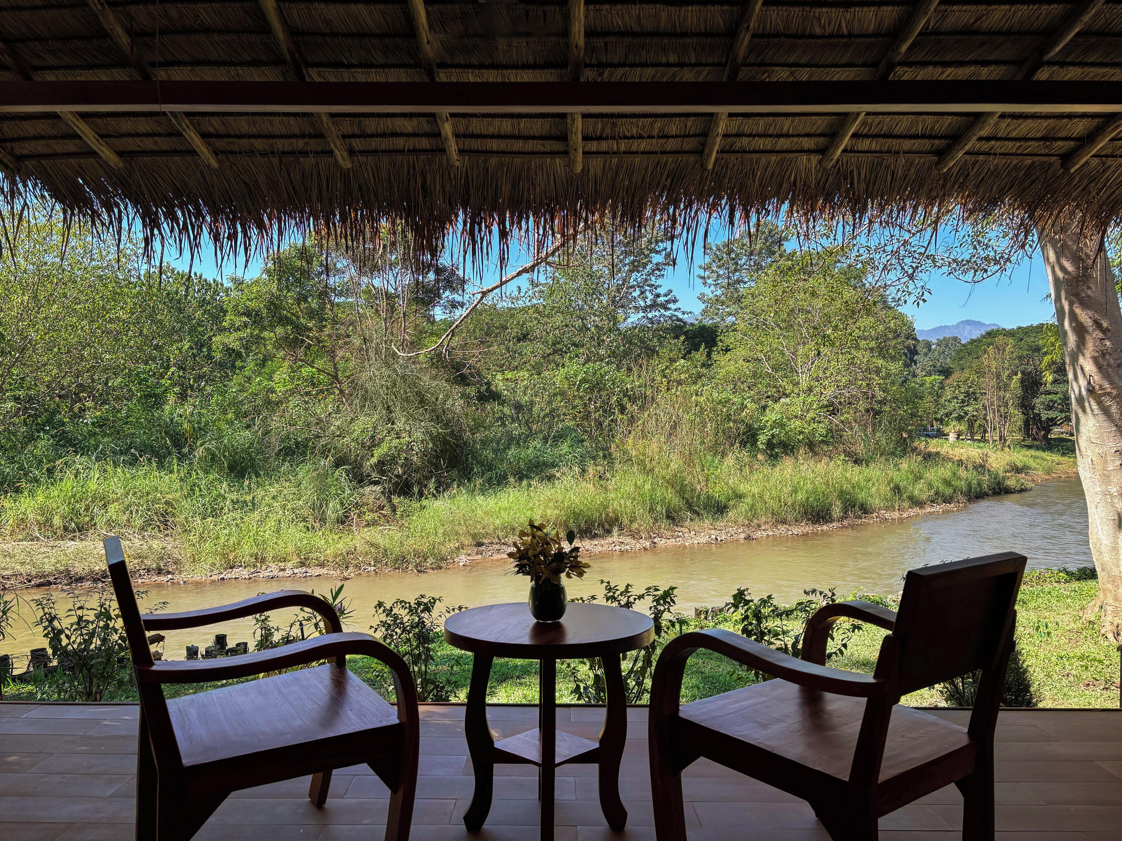 Ein Sitzbereich im Freien mit zwei Holzstühlen und einem kleinen Tisch, mit Blick auf einen Fluss und üppiges Grün.