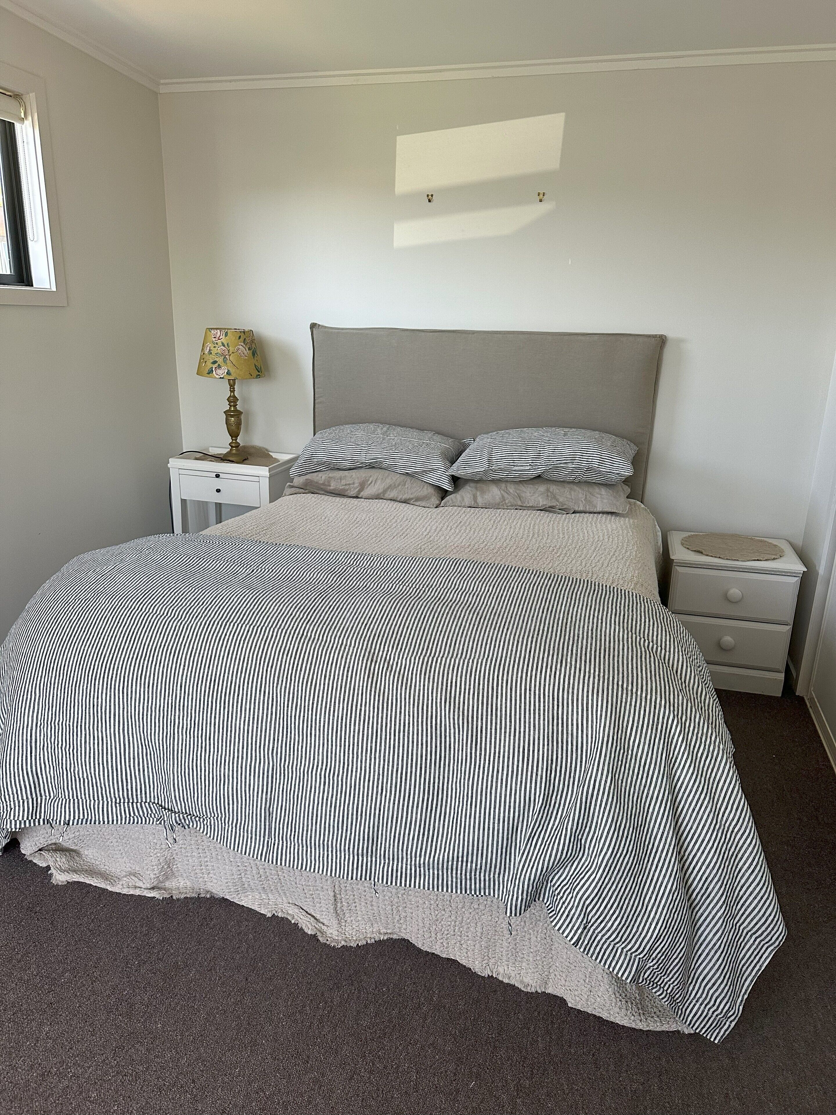 Cama bem arrumada com colcha listrada, duas almofadas, abajur e mesa de cabeceira com gavetas.