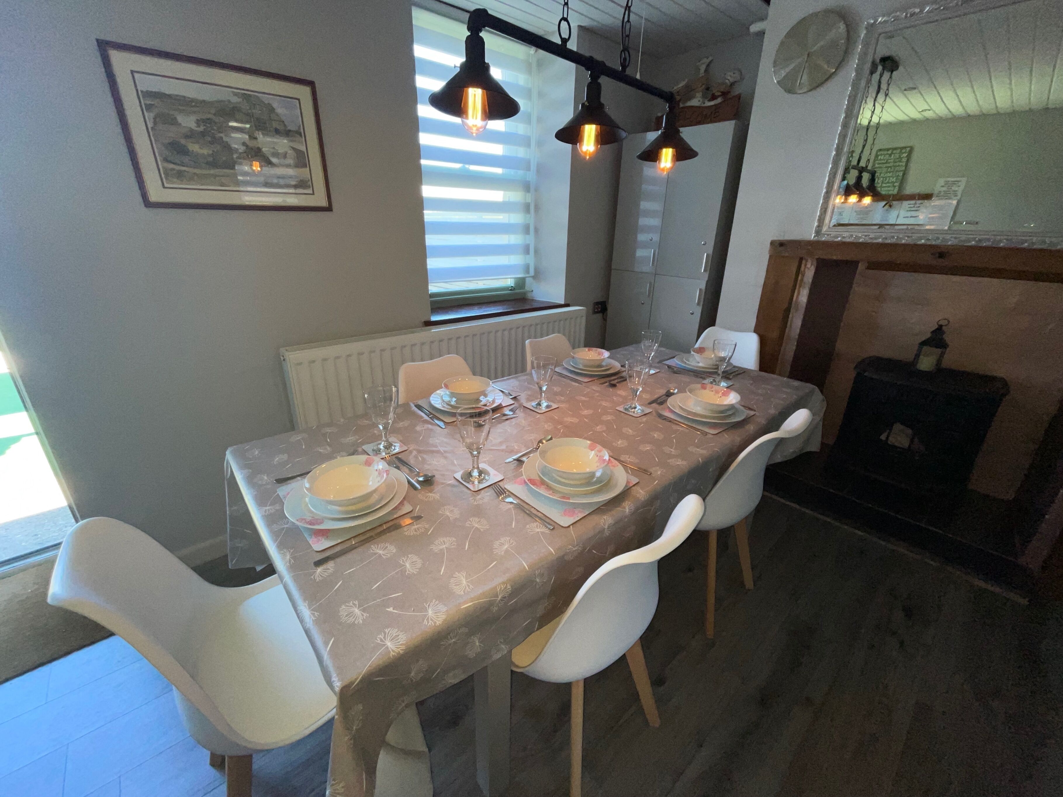 A dining table set for four with white chairs, a framed picture on the wall, and a window with blinds.