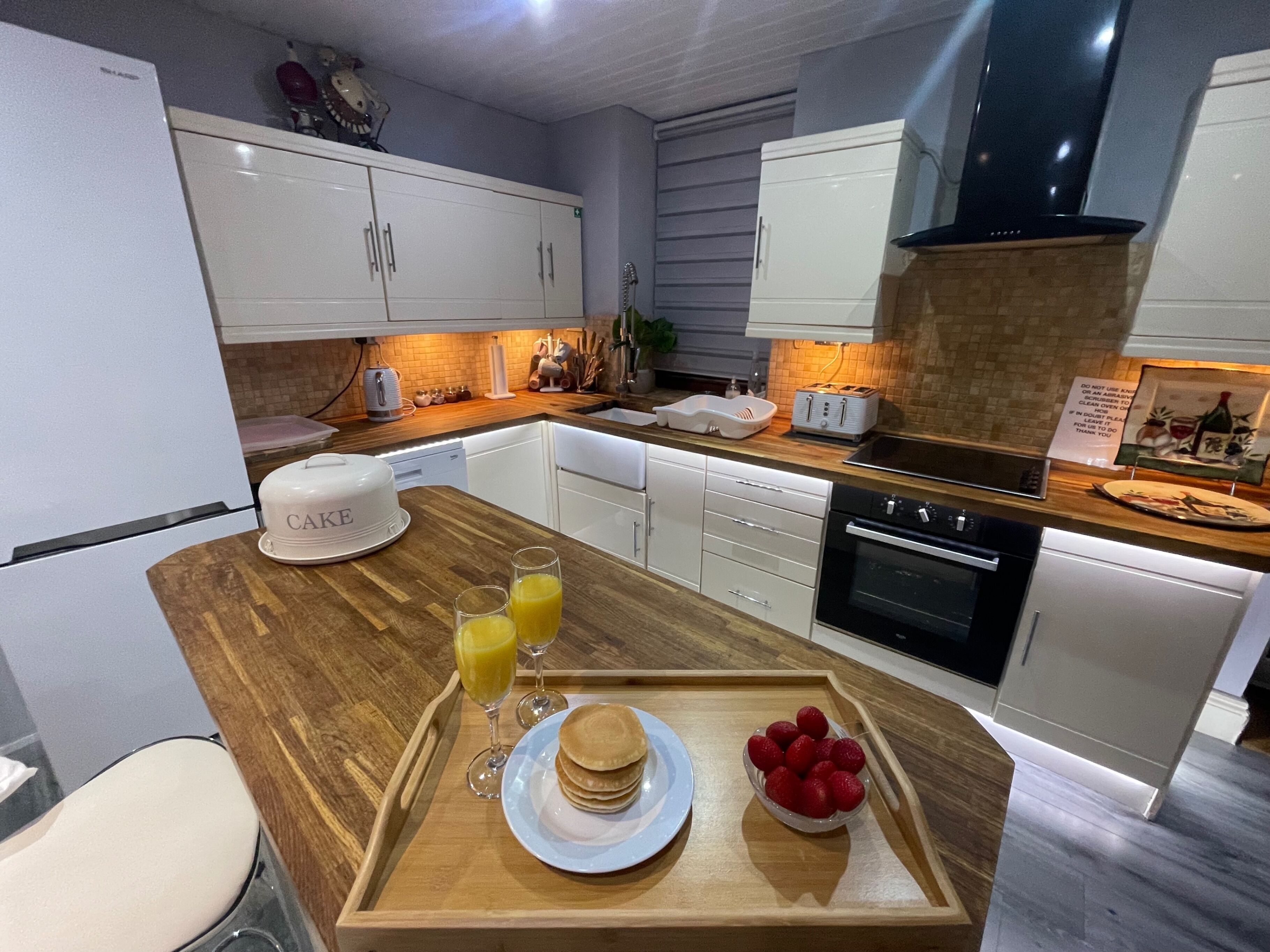 Une cuisine moderne avec des meubles blancs, un plan de travail en bois, un gâteau, des crêpes et un plateau de fraises.