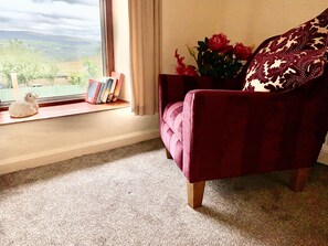 Ein Zimmer mit Fenster, einem roten Sessel, einer Blumenvase und einem kleinen Regal mit Büchern und einem Dekorationsgegenstand.