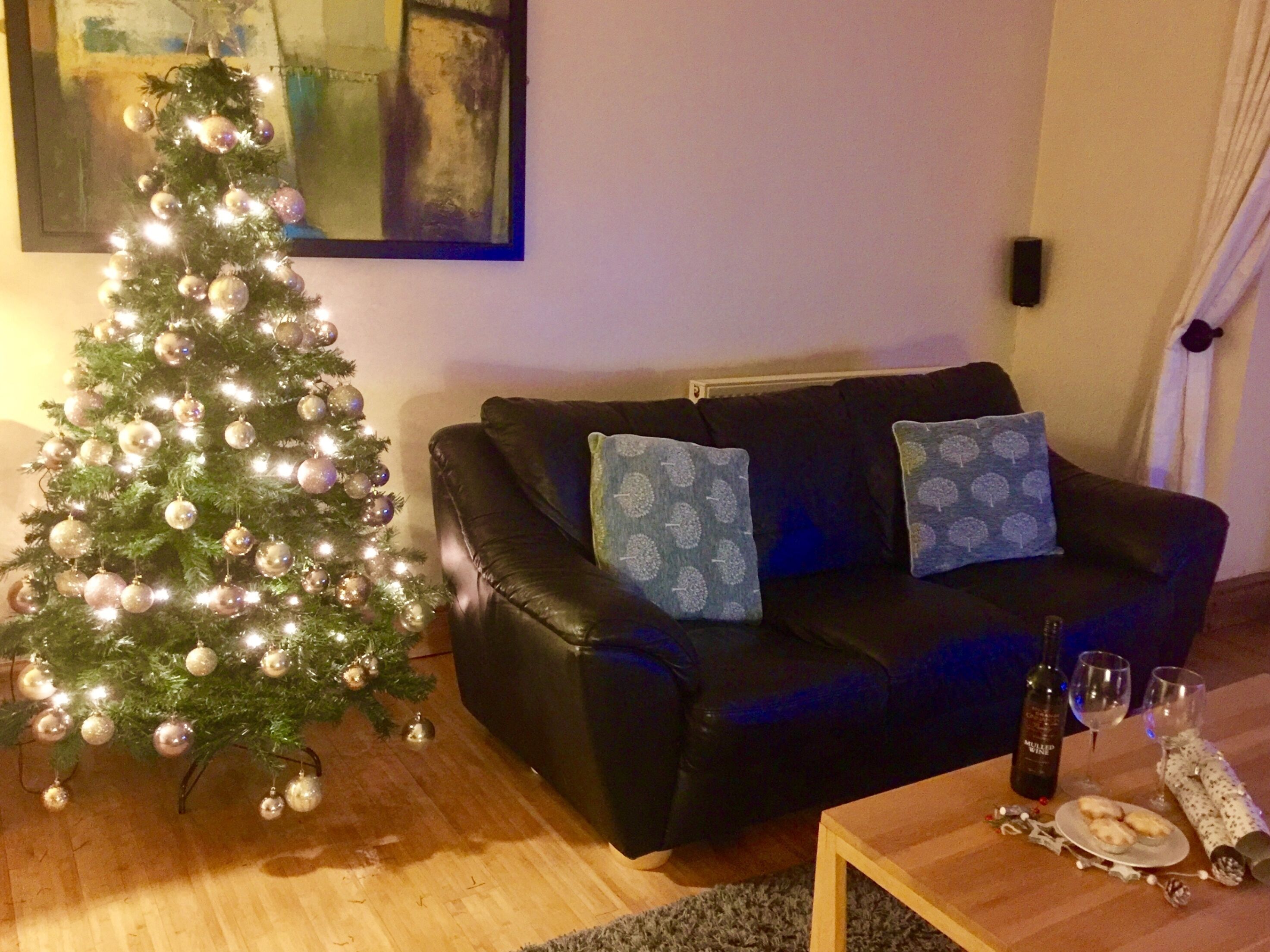 Een woonkamer met een kerstboom, een zwarte leren bank met gestreepte kussens, een houten salontafel met een bord koekjes en een fles wijn.