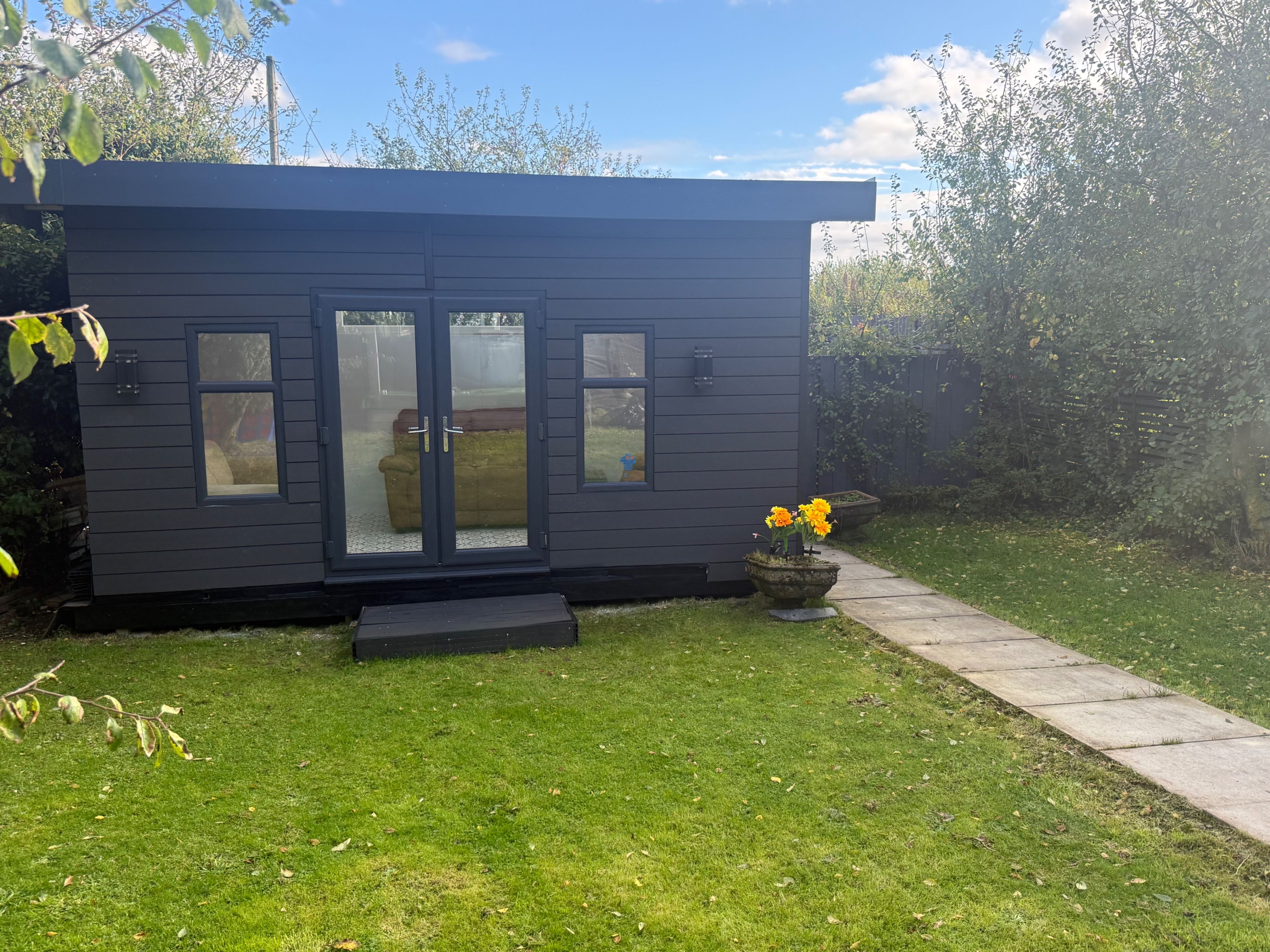 Une petite cabane de couleur sombre, dotée d’une porte coulissante en verre, d’un petit porche et d’une plante en pot ornée de fleurs jaunes.