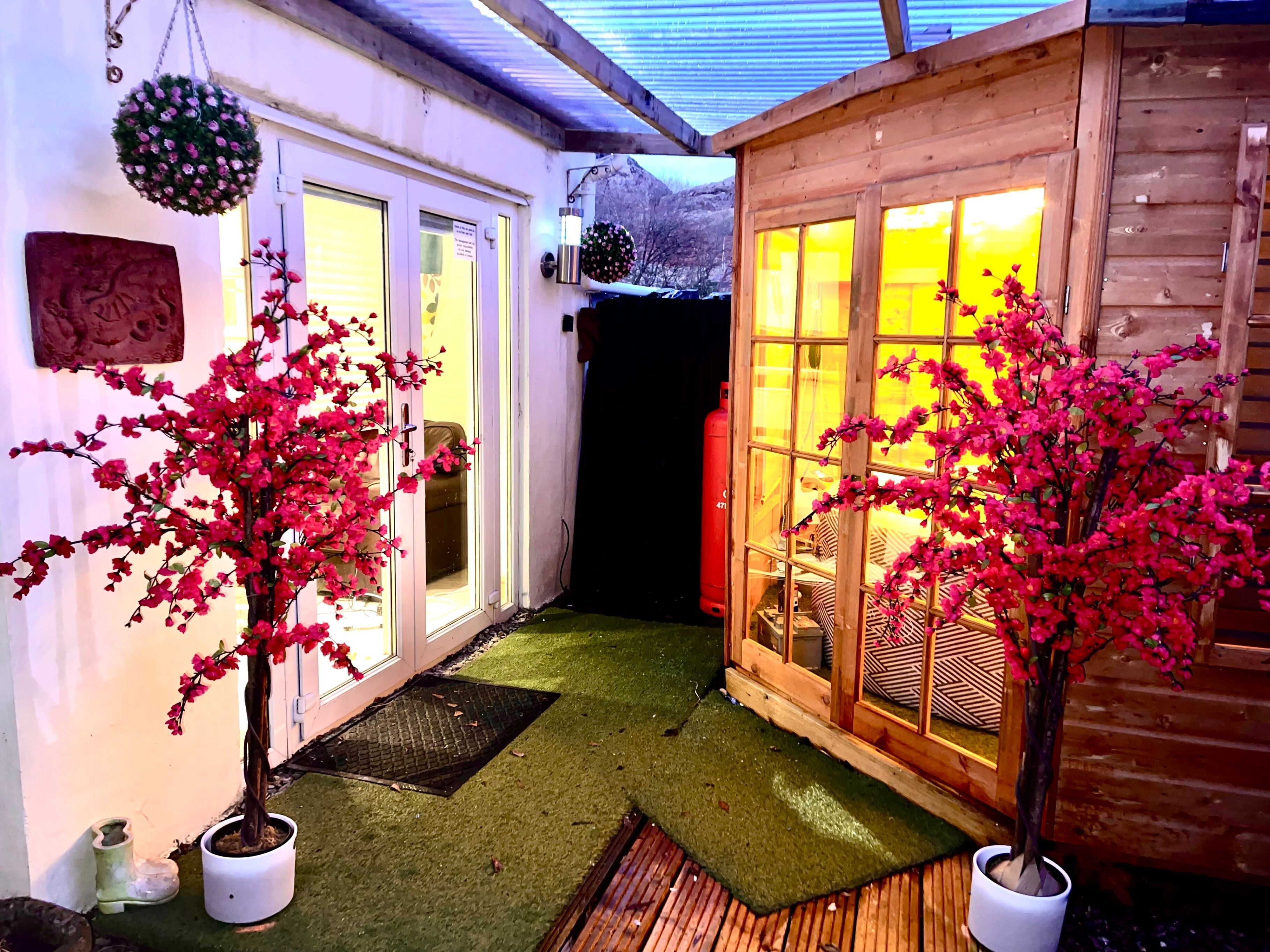 A wooden cabin with a glass door and windows, surrounded by potted plants and a small outdoor area with artificial grass.