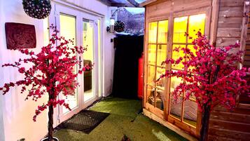 A wooden cabin with a glass door and windows, surrounded by potted plants and a small outdoor area with artificial grass.