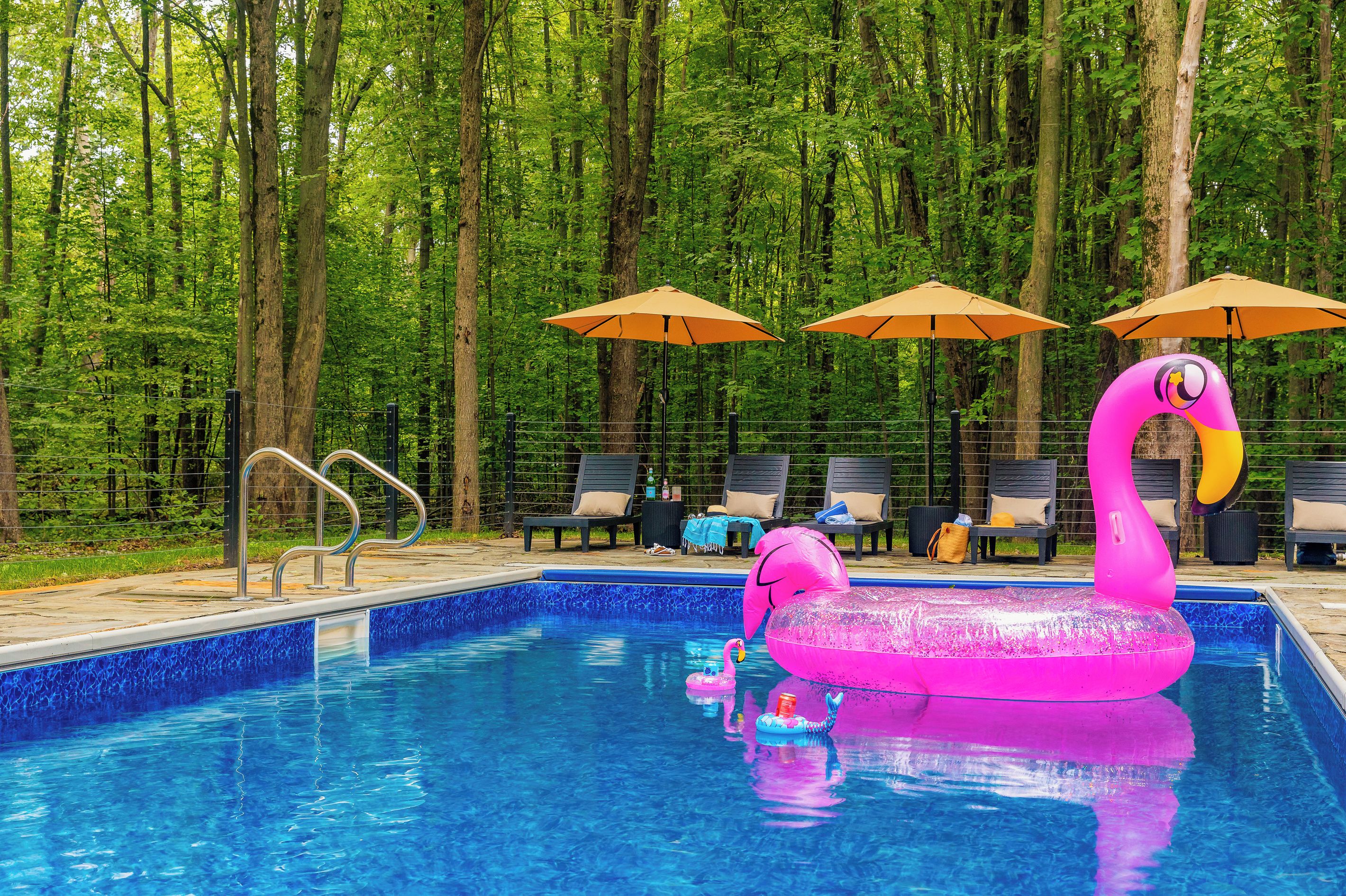 A swimming pool with a pink flamingo float, surrounded by lounge chairs and umbrellas, set against a forest backdrop.