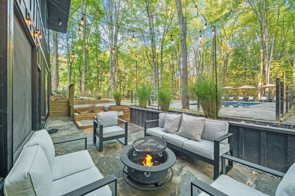 An outdoor patio with a fire pit, seating, and a view of a forest. - The Whispering Hollow – Private Pool, Hot Tub & Forest Escape Near Beach (Union Pier)