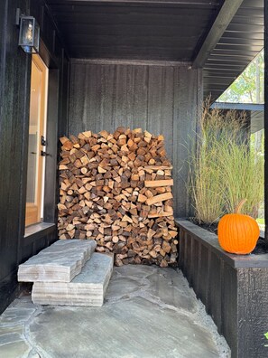 A stack of firewood against a dark wooden wall, a stone bench, and a carved pumpkin in a planter. - The Whispering Hollow – Private Pool, Hot Tub & Forest Escape Near Beach (Union Pier)