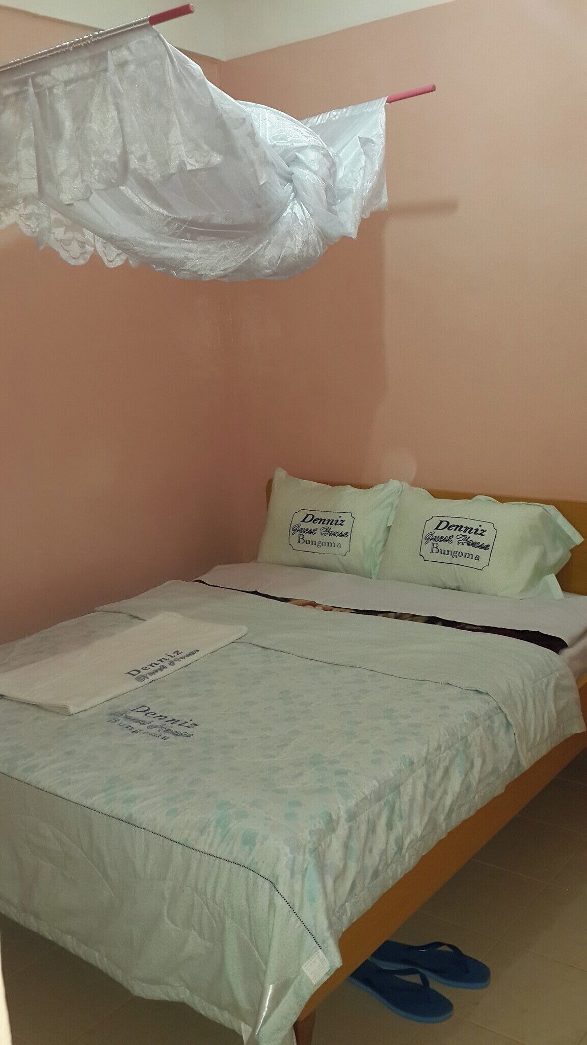 Una cama individual con sábana blanca y dos almohadas, una bordada con 'Dennis Guest House Bungoma'. Una bolsa de plástico cuelga del cabecero.