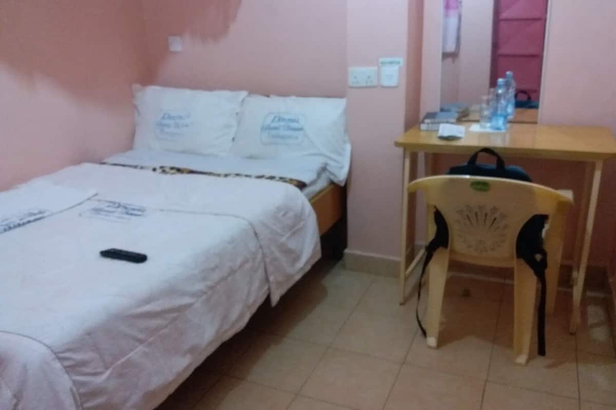 A small, single-bed room with a wooden desk and chair, a water bottle, and a remote control on the bed.