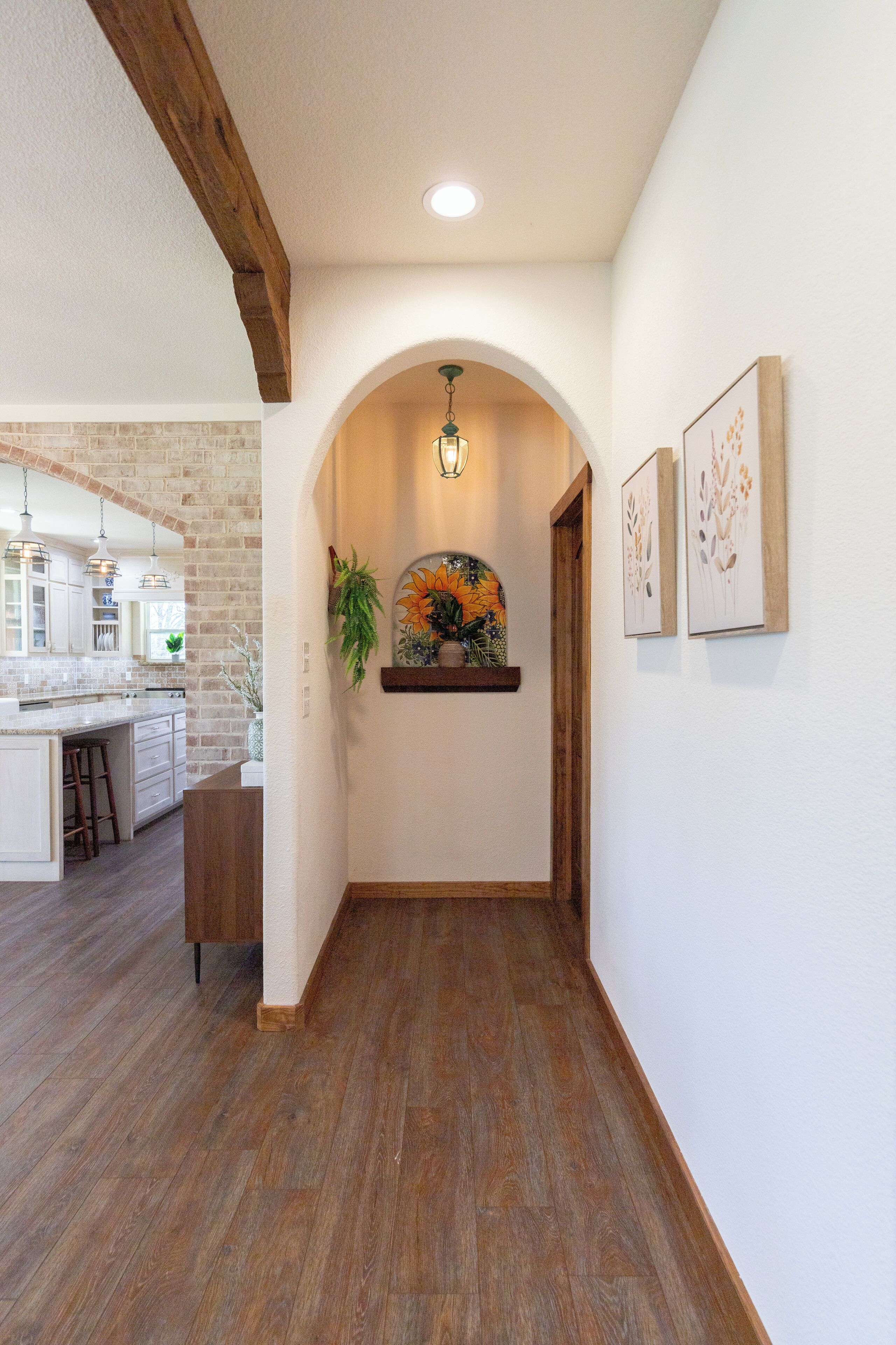 A hallway with wooden flooring, a brick wall, and a kitchen area visible through an archway.