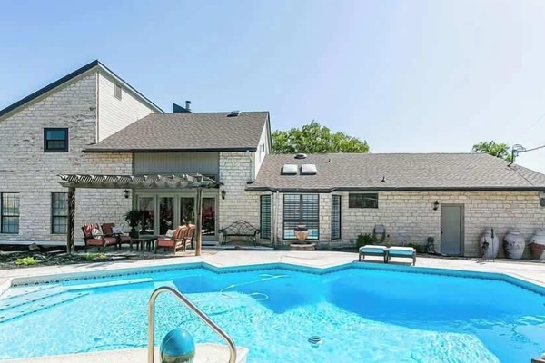 Backyard Poolside – A spacious view of the pool area showing the home’s rear stone exterior, pergola-covered patio seating, and large windows that open to the backyard.