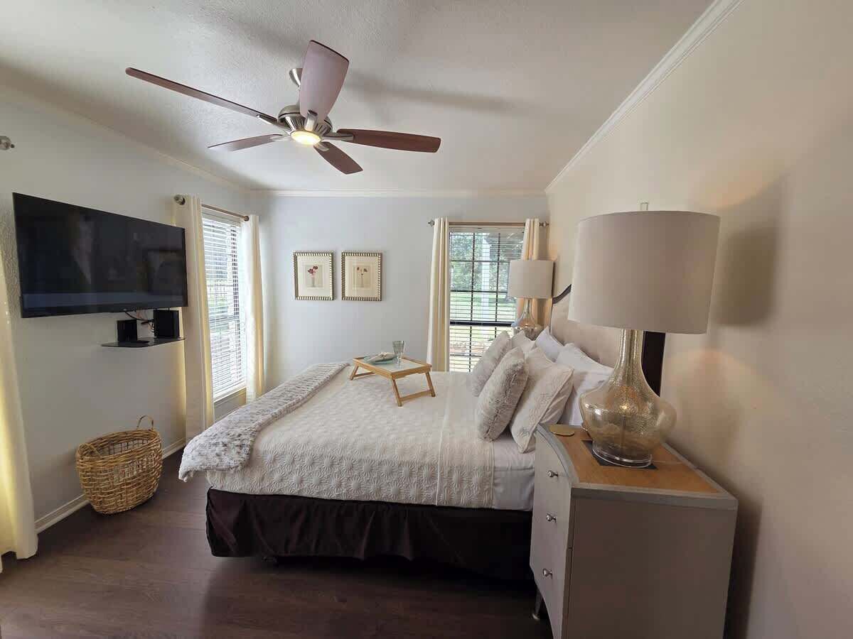 A bedroom with a bed, a ceiling fan, a TV, a bedside table with a lamp, and a window with blinds.