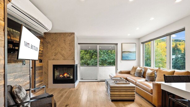 Een moderne woonkamer met een open haard, een bank, een eettafel en een groot raam met uitzicht op bomen.