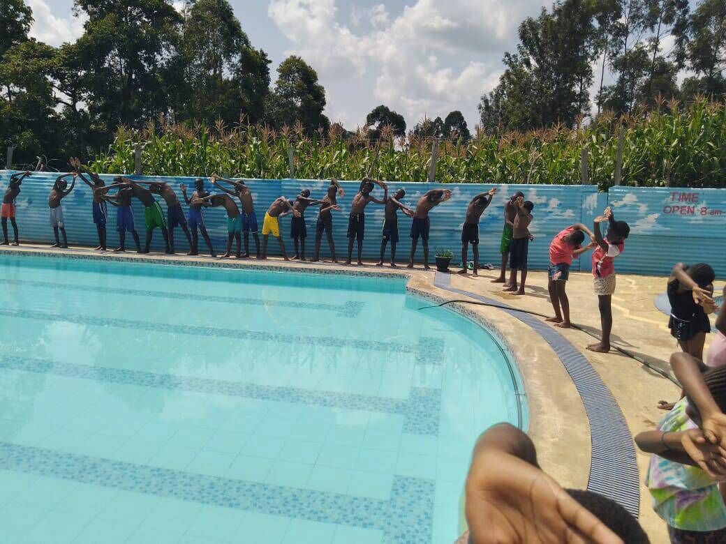 En swimmingpool med mennesker stående ved kanten, et blåt hegn og træer i baggrunden.