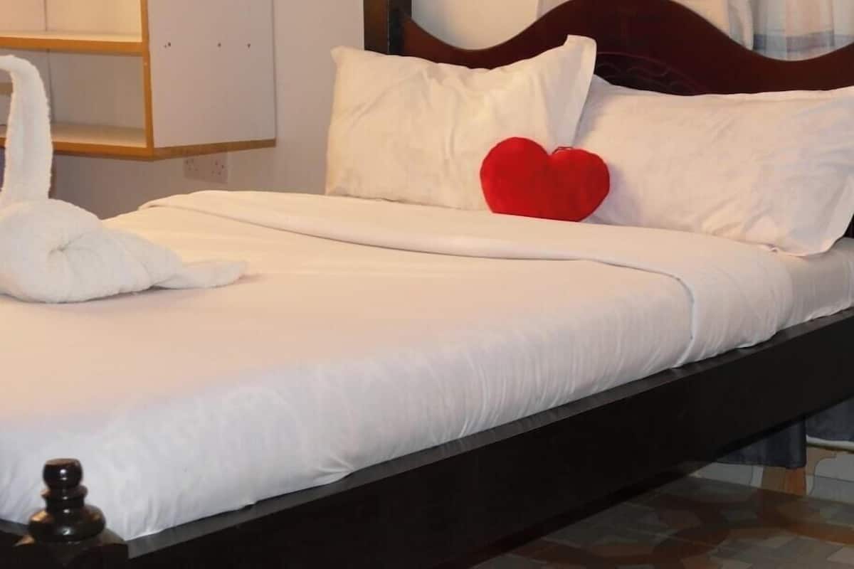 A bed with white bedding, a red heart pillow, and a fluffy pink rug on a patterned floor.