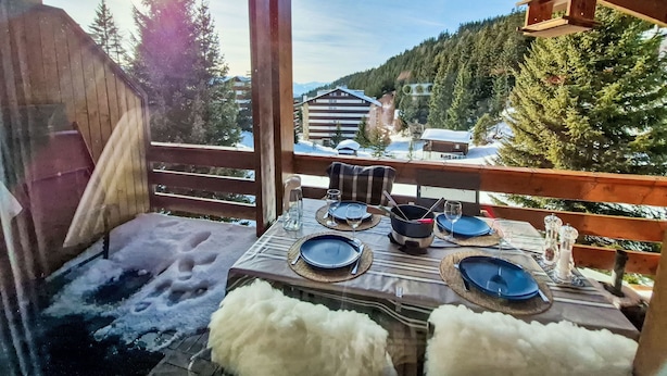 Een houten terras met een eettafel, met uitzicht op een besneeerd landschap met gebouwen en bomen.