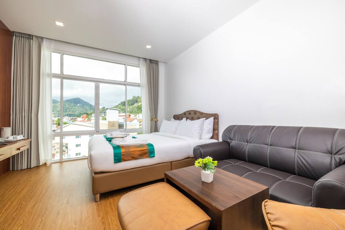A hotel room with a bed, a sofa, a small table, and a window with a view of buildings and hills.