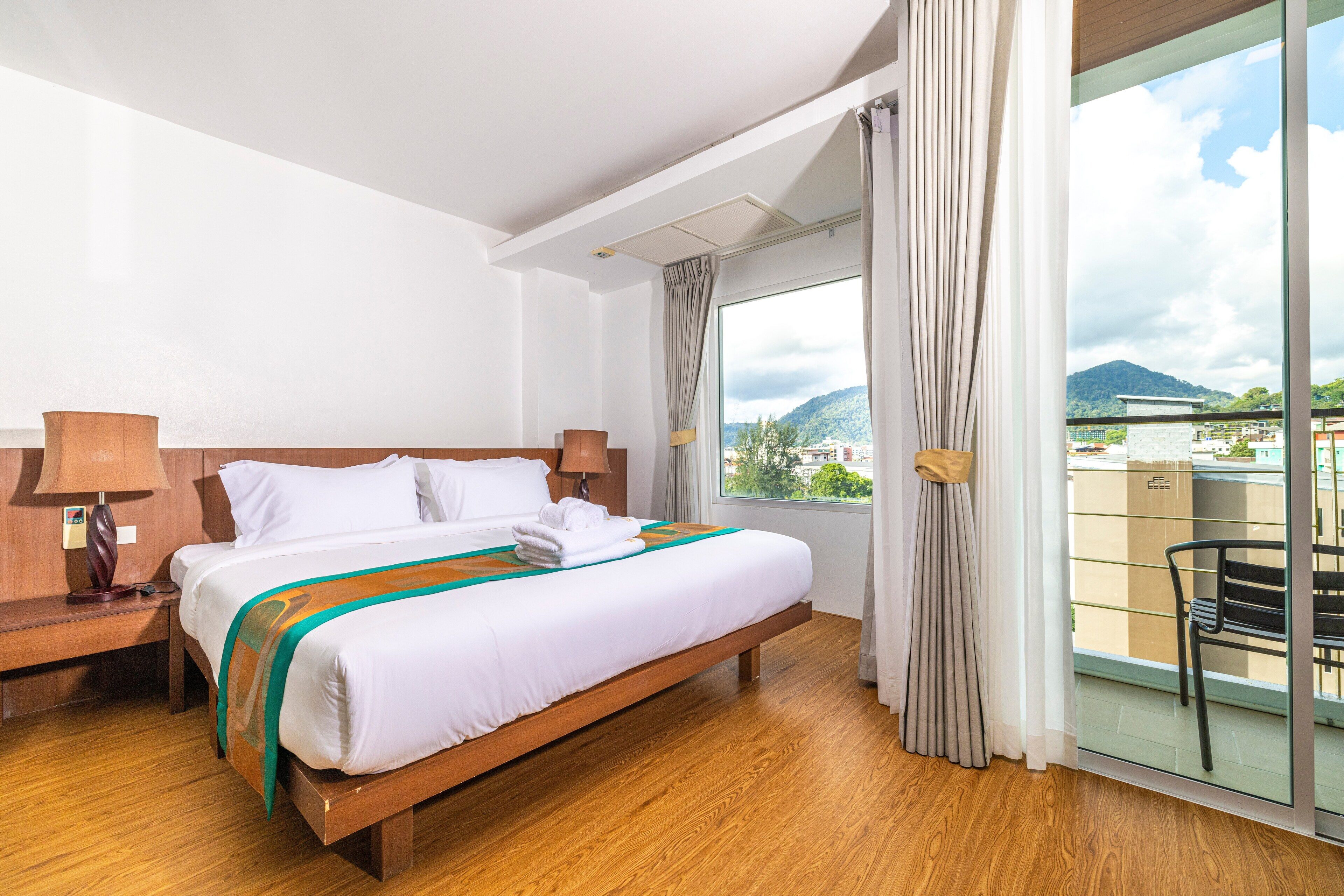 A hotel room with a wooden bed, a nightstand, a lamp, a window with curtains, and a view of a mountain and buildings.