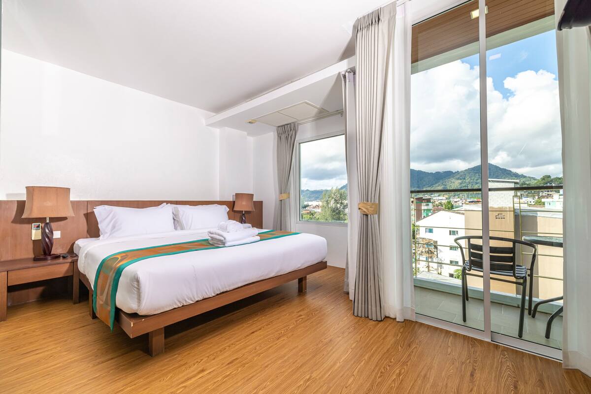 A hotel room with a large bed, wooden floor, a nightstand with a lamp, a window with curtains, and a view of buildings and mountains.