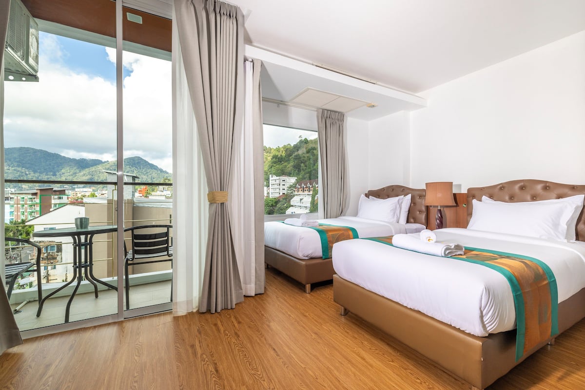 A hotel room with two beds, a balcony with a table and chairs, and a view of mountains and buildings.