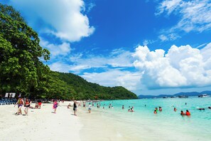 A beach with clear turquoise water, people swimming and sunbathing, and a forested hill in the background. - Aura Hotel Phuket (Patong)