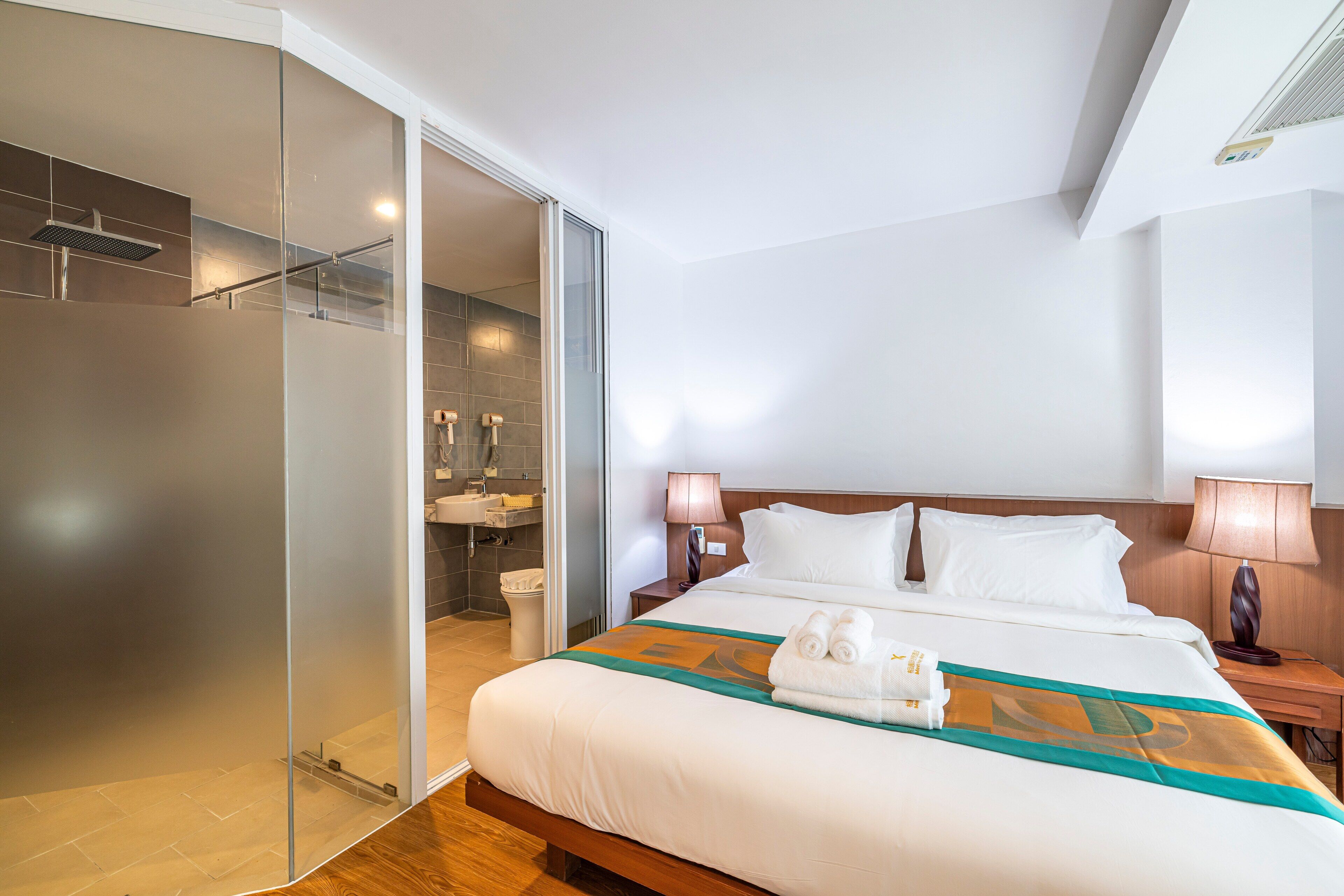 Quarto de hotel com cama, mesa de cabeceira, luminária, banheiro com chuveiro e porta de vidro que dá para o box.