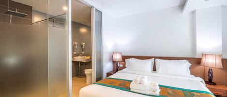 Quarto de hotel com cama, mesa de cabeceira, luminária, banheiro com chuveiro e porta de vidro que dá para o box.