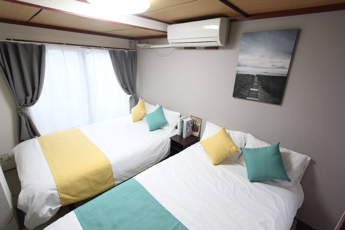 A hotel room with two beds, each with white bedding and teal and yellow accents. There is a window with grey curtains, an air conditioning unit mounted on the wall, and a framed picture above the beds.