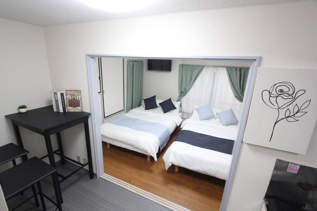 A small, compact room with two beds, a desk, a chair, a TV, and a refrigerator.